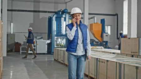 Photo for Beautiful Caucasian manager in helmet holding laptop while speaking with someone on smartphone. Young woman remotely talking with partners or clients while standing in manufacturing facility. - Royalty Free Image