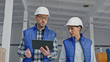 Photo for Camera view of two Caucasian workers actively talking with each other while walking on large storage or industrial factory. Handsome man telling something important while young woman disagreeing. - Royalty Free Image