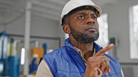 Photo for Close-up portrait of concentrated concerned hard-working thoughtful professional African-American specialist wearing helmet and vest verifying information from his device. - Royalty Free Image