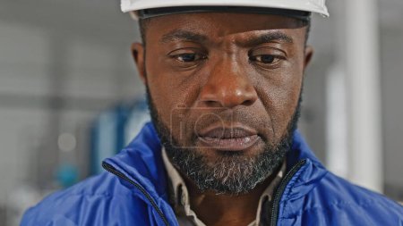 Photo for Close-up portrait of concentrated concerned hard-working thoughtful professional African-American specialist wearing helmet and vest verifying information from his device. - Royalty Free Image