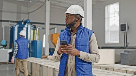 Photo for Zooming in on African American man wearing helmet while standing in middle of large facility. Specialist typing message on his phone. Considering production capacity while chatting with director. - Royalty Free Image