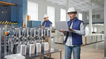 Photo for Caucasian female wearing vest and helmet for protection. Standing next to foil production line. Checking data while recording numbers on laptop. Man coming behind with digital device. - Royalty Free Image