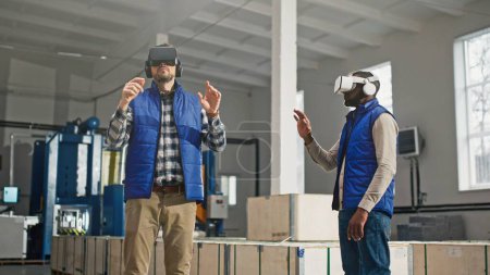 Téléchargez les photos : Gros plan de deux ingénieurs masculins d'âge moyen en casque et gilets bleus debout dans l'usine et ayant une merveilleuse expérience de réalité virtuelle. À l'intérieur. Les hommes en lunettes VR bougent leurs mains dans l'air. - en image libre de droit