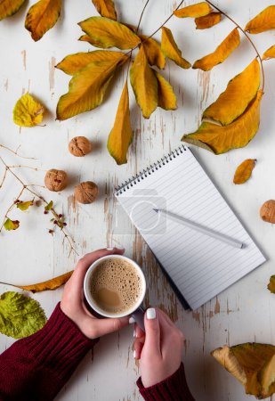 Photo for Top view of female hands holding cup of coffee on shabby table with yellow scattered leaves, walnuts and notepad with pen - Royalty Free Image