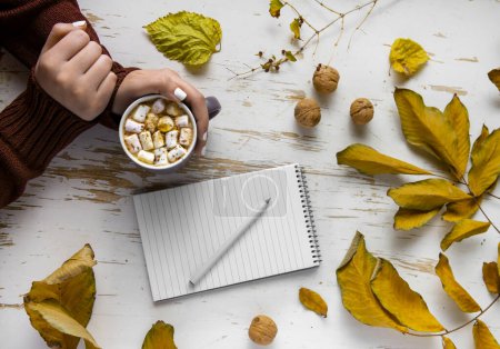 Photo for Top view of female hands holding cup of cocoa with marshmallow on shabby table with yellow scattered leaves, walnuts and notepad with pen, autumn composition - Royalty Free Image