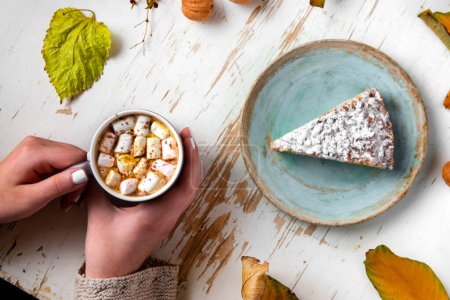 Photo for Top view of female hands holding marshmallows on shabby table with piece of cake, walnuts and scattered leaves, autumn composition - Royalty Free Image