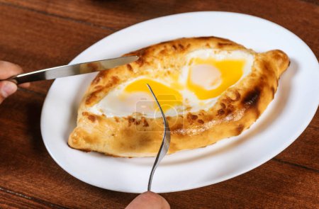 Foto de Hombre comiendo adjar Khachapuri. Pastel nacional georgiano khachapuri con huevo y queso en el plato blanco sobre fondo de madera. - Imagen libre de derechos