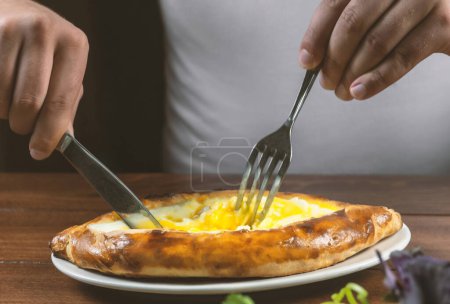 Téléchargez les photos : Homme mangeant adjar Khachapuri. Tarte nationale géorgienne khachapuri avec oeuf et fromage dans la plaque blanche sur fond de bois. - en image libre de droit