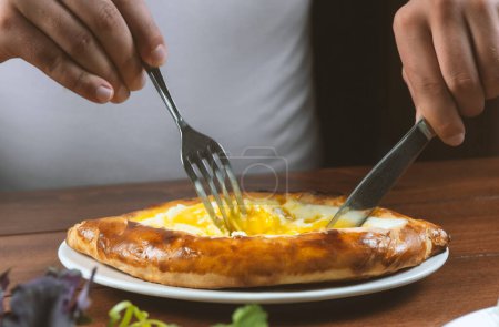 Téléchargez les photos : Un gaucher mangeant Adjar Khachapuri. Tarte nationale géorgienne khachapuri avec oeuf et fromage dans la plaque blanche sur fond de bois. - en image libre de droit