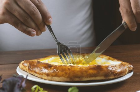 Foto de Hombre zurdo comiendo adjar Khachapuri. Pastel nacional georgiano khachapuri con huevo y queso en el plato blanco sobre fondo de madera. - Imagen libre de derechos