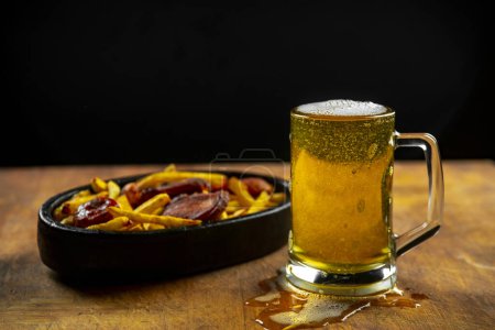 Foto de Espuma en un vaso con cerveza y salchichas fritas con patatas sobre un fondo de madera.Oktoberfest. - Imagen libre de derechos