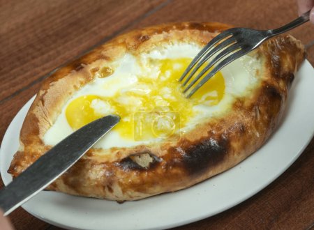 Photo for Man eating adjar Khachapuri. Georgian national pie khachapuri with egg and cheese in the white plate  on wooden background. - Royalty Free Image