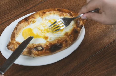 Photo for Man eating adjar Khachapuri. Georgian national pie khachapuri with egg and cheese in the white plate  on wooden background. - Royalty Free Image