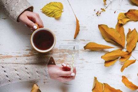 Photo for Top view of female hands holding cup of tea and thermometer on shabby table with yellow scattered leaves . autumn composition - Royalty Free Image