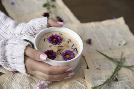 Photo for Female hands holding a cup of cappuccino with flowers on a book background - Royalty Free Image