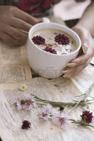 Téléchargez les photos : Mains féminines tenant une tasse de cappuccino avec des fleurs sur un fond de livre - en image libre de droit