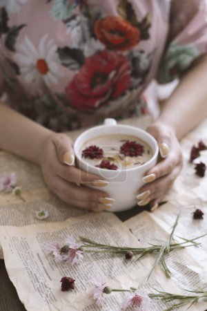 Téléchargez les photos : Mains féminines tenant une tasse de cappuccino avec des fleurs sur un fond de livre - en image libre de droit