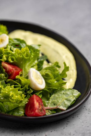 Foto de Fresh green salad with quail eggs, cherry tomatoes and guacamole in black plate on grey background - Imagen libre de derechos