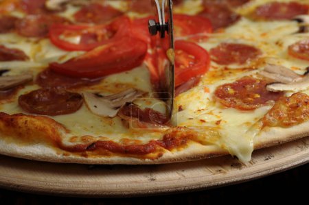 Foto de Proceso de corte de pizza con jamón, queso y tomates en una tabla de madera. - Imagen libre de derechos