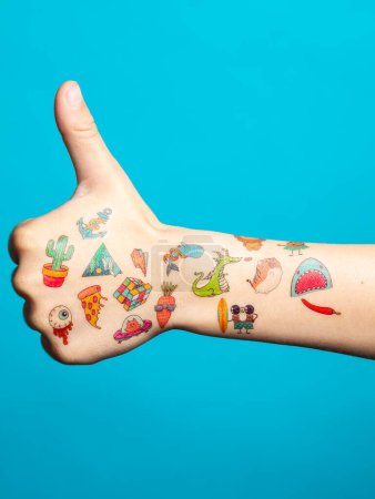 Téléchargez les photos : Pouce levée de la main d'un petit garçon avec beaucoup de tatouages multicolores de transfert sur un fond bleu isolé - en image libre de droit