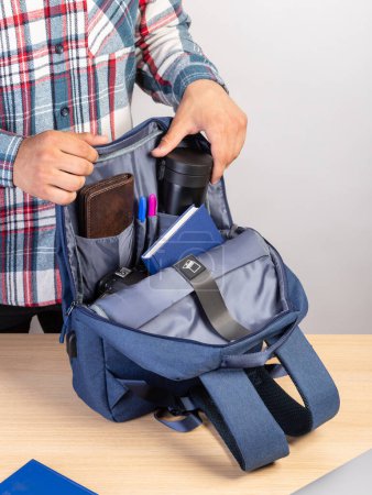 Foto de Primer plano de un joven estudiante sacando una taza de café de una mochila que también contiene una computadora portátil, billetera, bloc de notas - Imagen libre de derechos