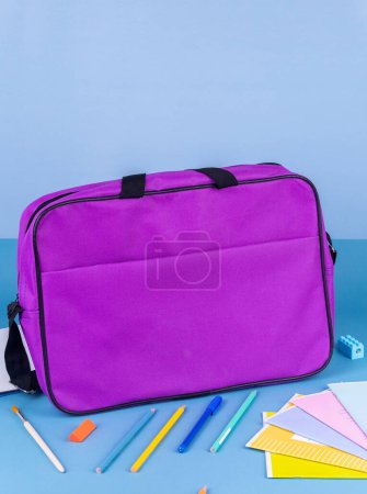 Foto de Carpeta de maletín púrpura para documentos y artículos escolares en una mesa azul brillante junto a artículos de papelería - Imagen libre de derechos