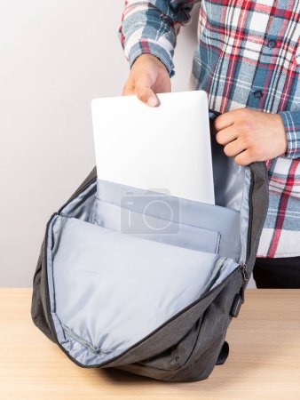 Foto de Un hombre pone un portátil en una mochila afuera, sobre un fondo claro. Un joven mete un portátil en una mochila. Un hombre saca un portátil de su mochila. - Imagen libre de derechos