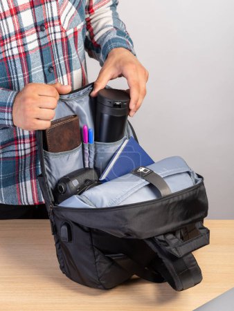 Téléchargez les photos : Gros plan d'un jeune étudiant qui sort une tasse à café d'un sac à dos qui contient également un ordinateur portable, un portefeuille, un bloc-notes - en image libre de droit