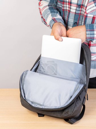 Téléchargez les photos : Un homme met un ordinateur portable dans un sac à dos à l'extérieur, sur un fond clair. Un jeune homme met un ordinateur portable dans un sac à dos. Un homme sort un ordinateur portable de son sac à dos. - en image libre de droit
