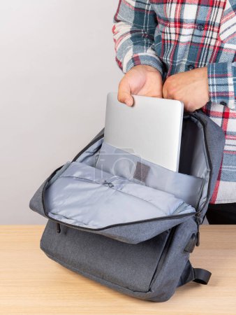 Foto de Un hombre con una camisa a cuadros pone su mano en un portátil gris en una elegante mochila gris. - Imagen libre de derechos