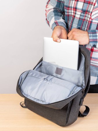 Photo for The hands of an unrecognizably cropped man in a plaid shirt put a laptop in a backpack, going to work. - Royalty Free Image