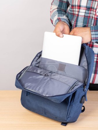 Téléchargez les photos : Un homme met un ordinateur portable dans un sac à dos à l'extérieur, sur un fond clair. Un jeune homme met un ordinateur portable dans un sac à dos. Un homme sort un ordinateur portable de son sac à dos. - en image libre de droit