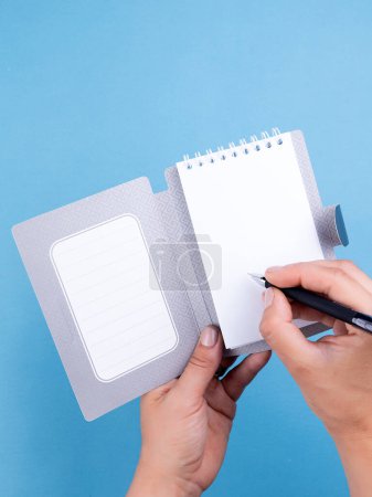 Photo for Design concept - top view of a woman writing in a notebook with white sheets, bookmarks, a binder and a pen on a blue background for a layout. - Royalty Free Image
