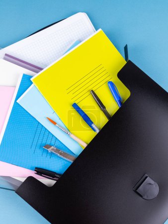 Photo for Black plastic  briefcase folder for documents and school items on a bright blue table next to stationery - Royalty Free Image