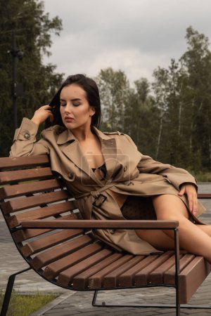 Photo for Outdoor closeup portrait of young beautiful woman in beige trench coat and black lingerie posing on bench against park . Fashion concept. - Royalty Free Image