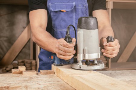 Téléchargez les photos : Jeune homme par profession charpentier constructeur égale une fraiseuse en bois sur une table en bois dans l'atelier - en image libre de droit