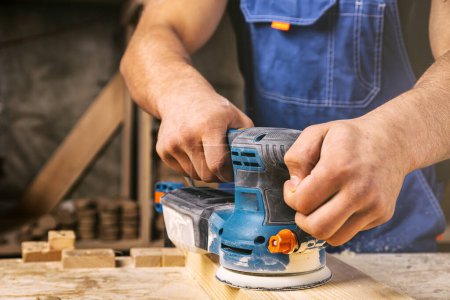 Photo for Close up of an young man builder carpenter equals polishes wooden board with a  random orbit sander  in the workshop - Royalty Free Image