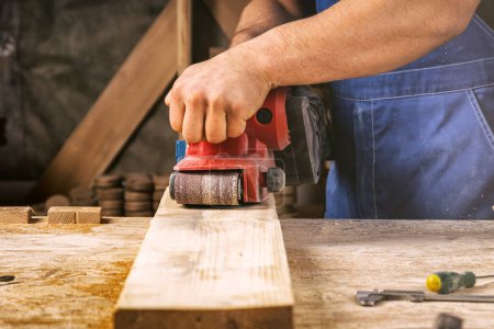 Téléchargez les photos : Gros plan d'un jeune homme constructeur charpentier égale polis planche en bois dans l'atelier - en image libre de droit