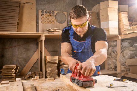 Téléchargez les photos : Portrait d'un jeune menuisier utilisant une ponceuse électrique pour polir un atelier de barin en bois - en image libre de droit