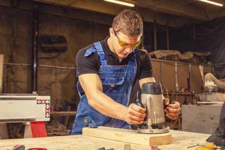 Téléchargez les photos : Un jeune homme portant des lunettes de sécurité menuisier prépare les bords de la planche de bois avec une fraiseuse dans l'atelier, sciure de bois vole sur les côtés - en image libre de droit