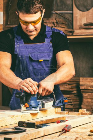 Téléchargez les photos : Jeune homme charpentier broyage du bois avec du papier de verre en menuiserie ou atelier de bricolage. Ponceuse électrique travaillant dans la menuiserie - en image libre de droit