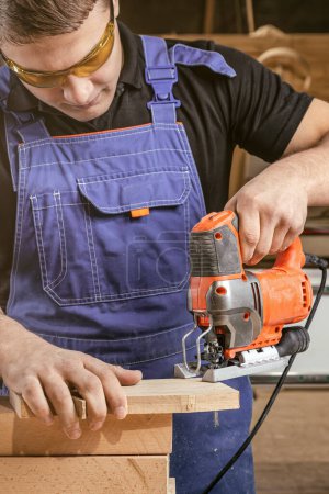 Photo for Close up of experienced carpenter in work clothes and small buiness owner  carpenter saw and processes the edges of a wooden bar with a jig saw  in a workshop - Royalty Free Image