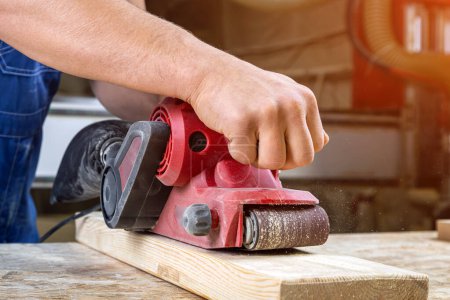 Photo for Young man builder carpenter equals polishes wooden board with a  random orbit sander  in the workshop - Royalty Free Image