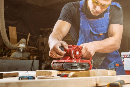 Téléchargez les photos : Jeune homme constructeur charpentier égale polis planche en bois avec une ponceuse d'orbite aléatoire dans l'atelier - en image libre de droit