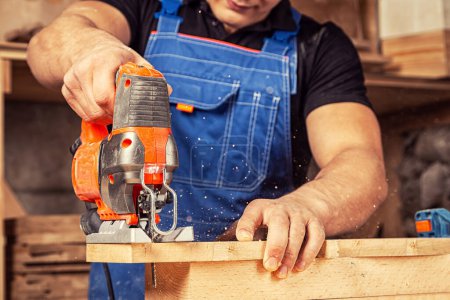 Photo for Close up of experienced carpenter in work clothes and small buiness owner  carpenter saw and processes the edges of a wooden bar with a jig saw  in a workshop - Royalty Free Image