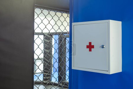 Téléchargez les photos : Gros plan d'une trousse de premiers soins vide en métal blanc avec une croix rouge accrochée sur un fond mural bleu. - en image libre de droit