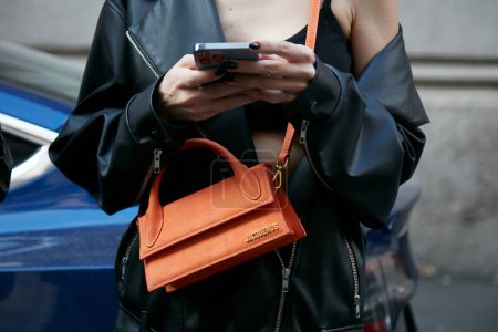 Foto de MILÁN, ITALIA - 21 DE SEPTIEMBRE DE 2022: Mujer con bolso Jacquemus de cuero naranja mirando el teléfono inteligente antes del desfile de moda Alberta Ferretti, Milan Fashion Week street style - Imagen libre de derechos
