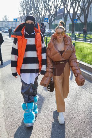 Téléchargez les photos : MILAN, ITALY - JANUARY 14, 2023: Man with black balaclava and orange jacket and woman with beige padded jacket and trousers before Emporio Armani fashion show, Milan Fashion Week street style - en image libre de droit