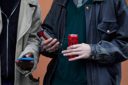 Foto de MILAN, ITALY - JANUARY 15, 2023: Men with Coca Cola can looking at smartphone before Etro fashion show, Milan Fashion Week street style - Imagen libre de derechos