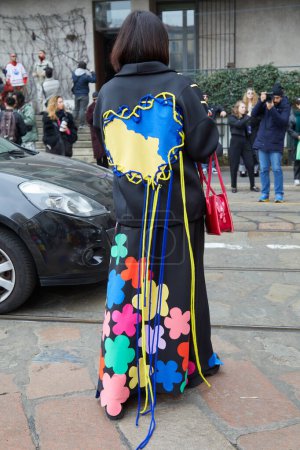Foto de MILÁN, ITALIA - 24 DE FEBRERO DE 2023: Mujer con abrigo negro con corazón amarillo y azul con cuerdas antes del desfile de moda Sportmax, Milan Fashion Week street style - Imagen libre de derechos